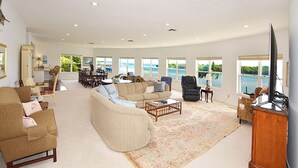 A spacious living room with a large sofa, a coffee table, and a television. There are multiple windows providing views of a body of water. - Sandman's Haven Retreat (Stuart)