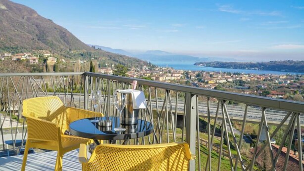 A balcony with yellow chairs and a table, overlooking a coastal town and mountains. - Hotel Vita (Salò)