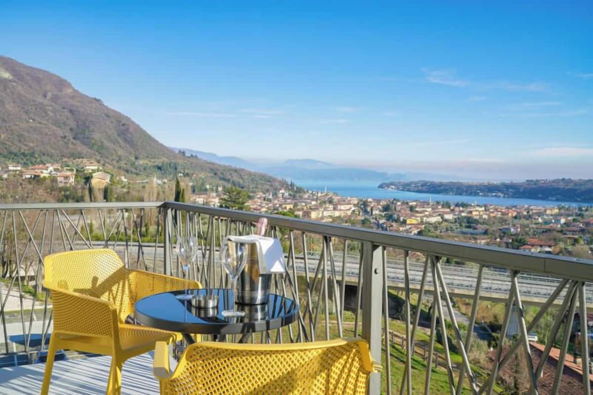 A balcony with yellow chairs and a table, overlooking a coastal town and mountains.