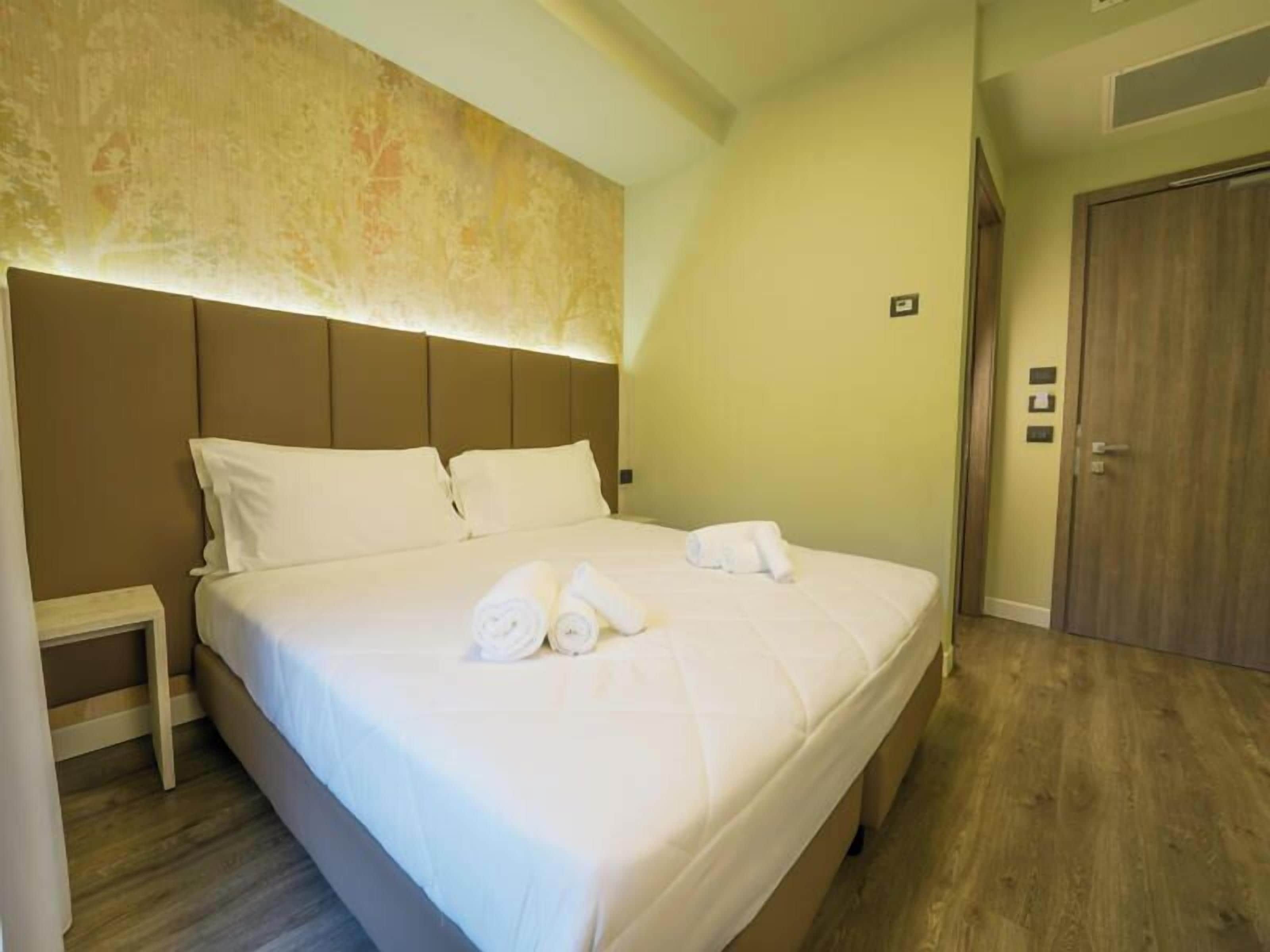 A neatly made bed with white linens and rolled towels, a bedside table, and a wooden door in a hotel room.