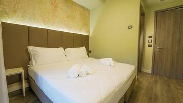 A neatly made bed with white linens and rolled towels, a bedside table, and a wooden door in a hotel room.