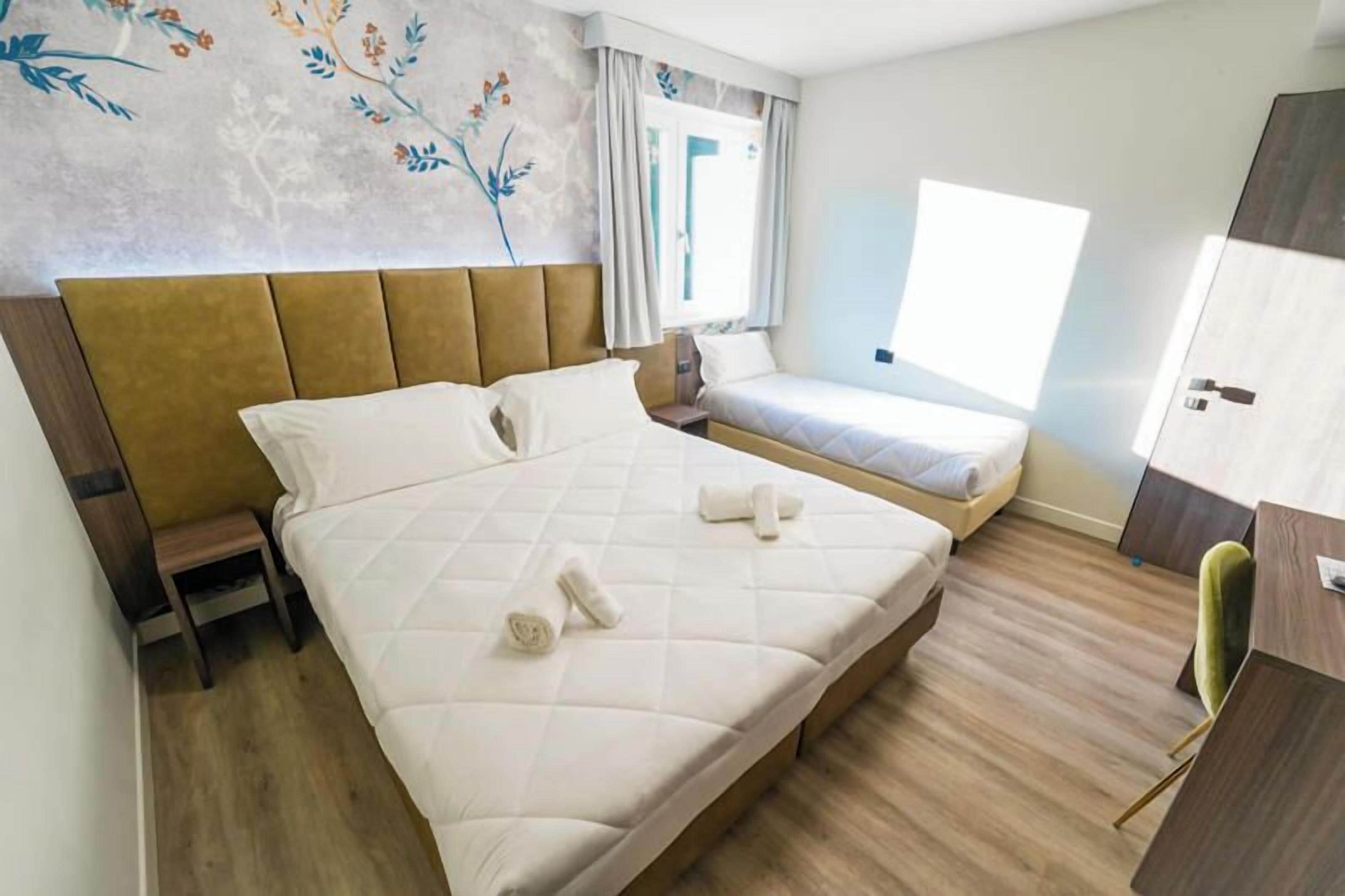 Hotellihuone, jossa on suuri sänky, pienempi sänky, työpöytä ja vaatekaappi.