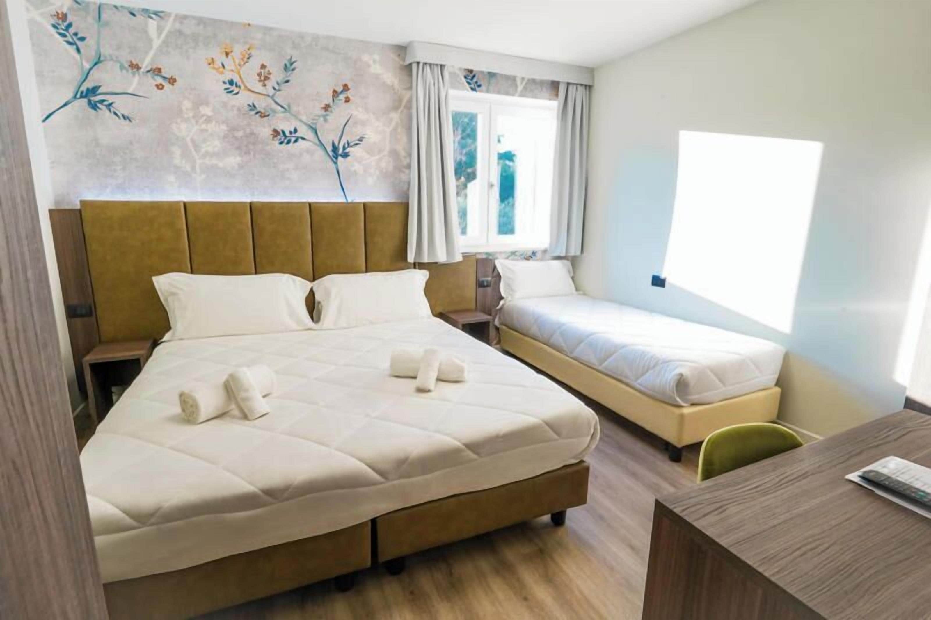 Ett hotellrum med en stor säng, en mindre säng, ett skrivbord och ett fönster med gardiner.