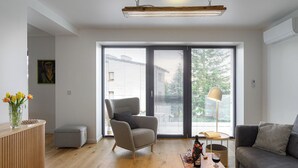 Una sala de estar moderna con un sillón gris, un sofá, una mesa de centro de madera con un libro y una botella, y una lámpara de pie.