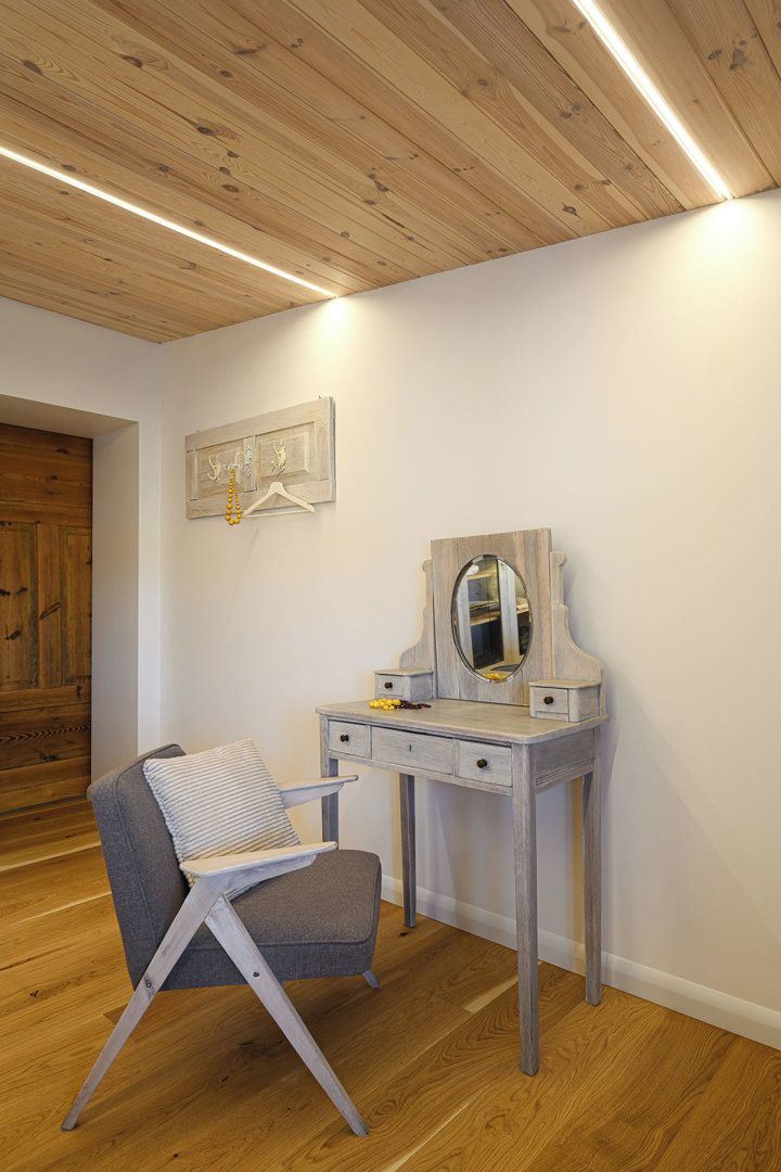 A room with a wooden ceiling, a grey chair, a small vanity with a mirror, and a wooden door.