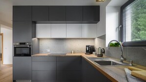 Una cocina moderna con armarios oscuros, encimera de madera, horno empotrado, cafetera y ventana con persianas.