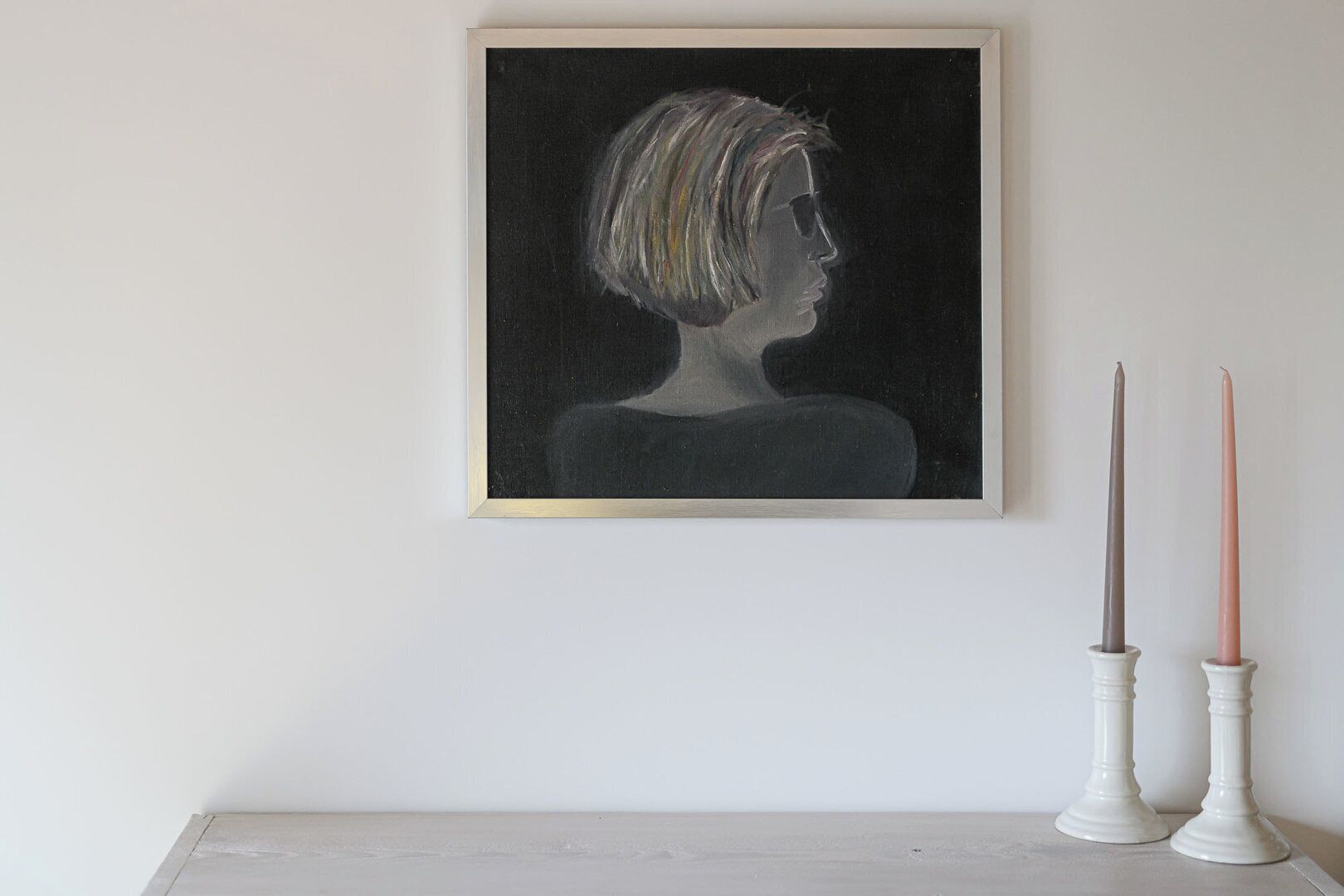 A framed painting of a person with short hair on a white wall, next to two white candle holders with candles.