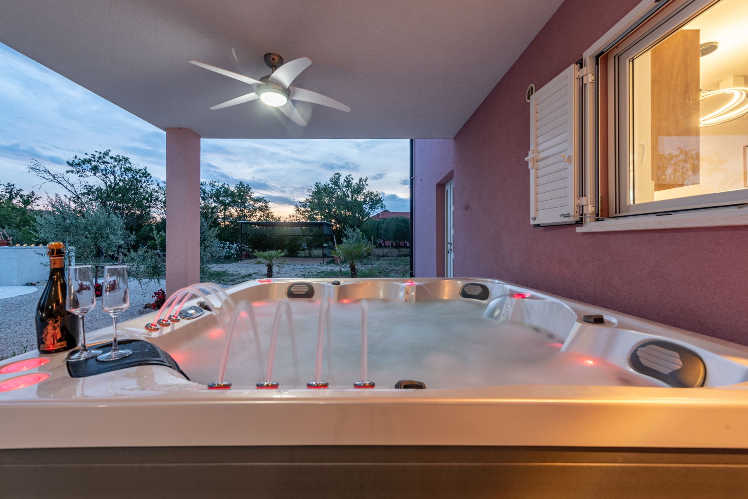 A hot tub with a ceiling fan, a bottle of wine, and glasses on a table.