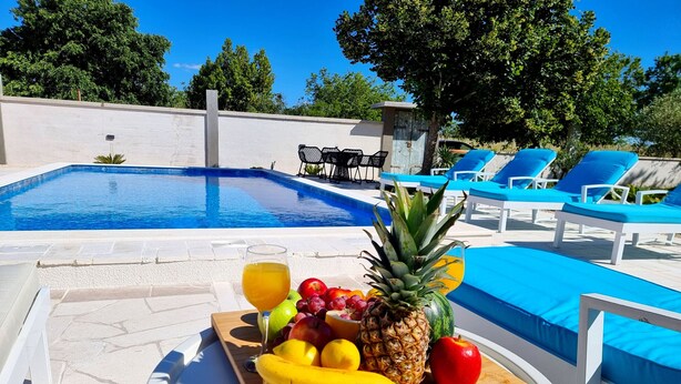 A poolside table with a fruit platter, a glass of juice, and a pool with blue lounge chairs. - Villa Rosea Domus Zadarvillas (Benkovac)