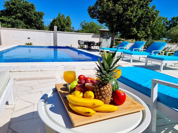 Una mesa junto a la piscina con una bandeja de frutas, un vaso de jugo y una piscina con sillas reclinables azules.