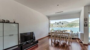 Ein geräumiges Wohnzimmer mit großem Fernseher, Holzboden, einem Essbereich mit Stühlen und Blick auf einen See und Hügel.