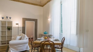 Ein Essbereich mit einem Tisch, gedeckt für vier Personen, umgeben von Holzstühlen. Eine weiße Couch steht an der Wand, und links ist ein Bücherregal zu sehen.