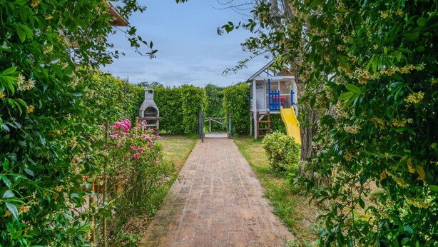 A brick pathway leading to a wooden playhouse with a slide, surrounded by lush greenery and flowering plants.