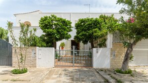 Un bâtiment blanc avec une entrée fermée par un portail, entouré de verdure et agrémenté d’un petit jardin.