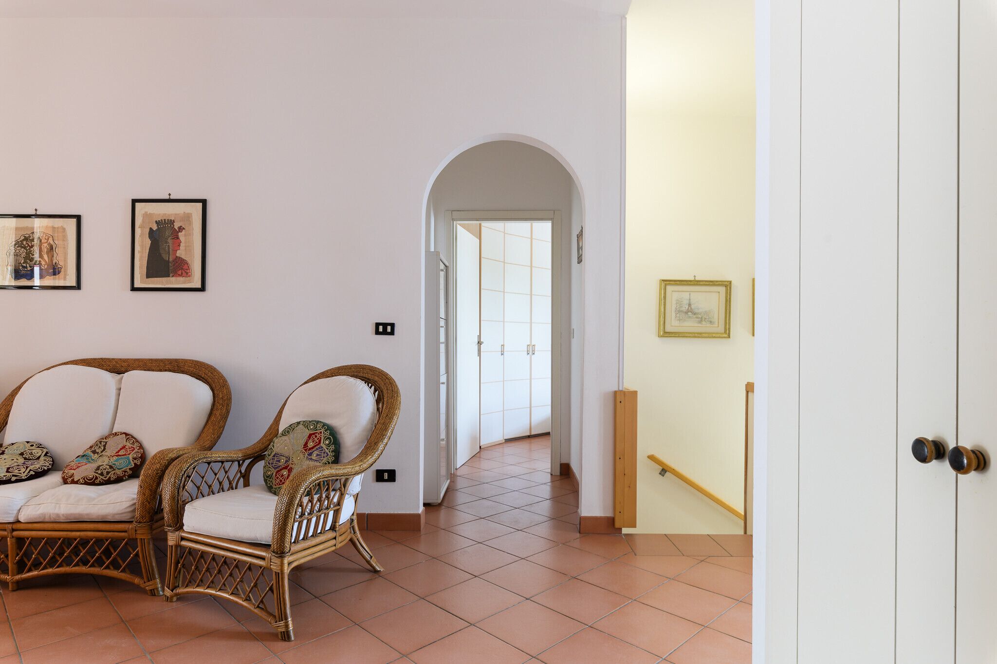 Una habitación con sillas de mimbre, una puerta blanca y un pasillo que conduce a otra estancia.