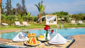 A dining table with a meal set, a pool, and a tropical garden in the background. - Villa Dar Minzah (Aghouatim)