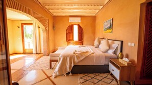 A bedroom with a large bed, a wooden nightstand, a small table, and a painting on the wall. - Villa Dar Minzah (Aghouatim)