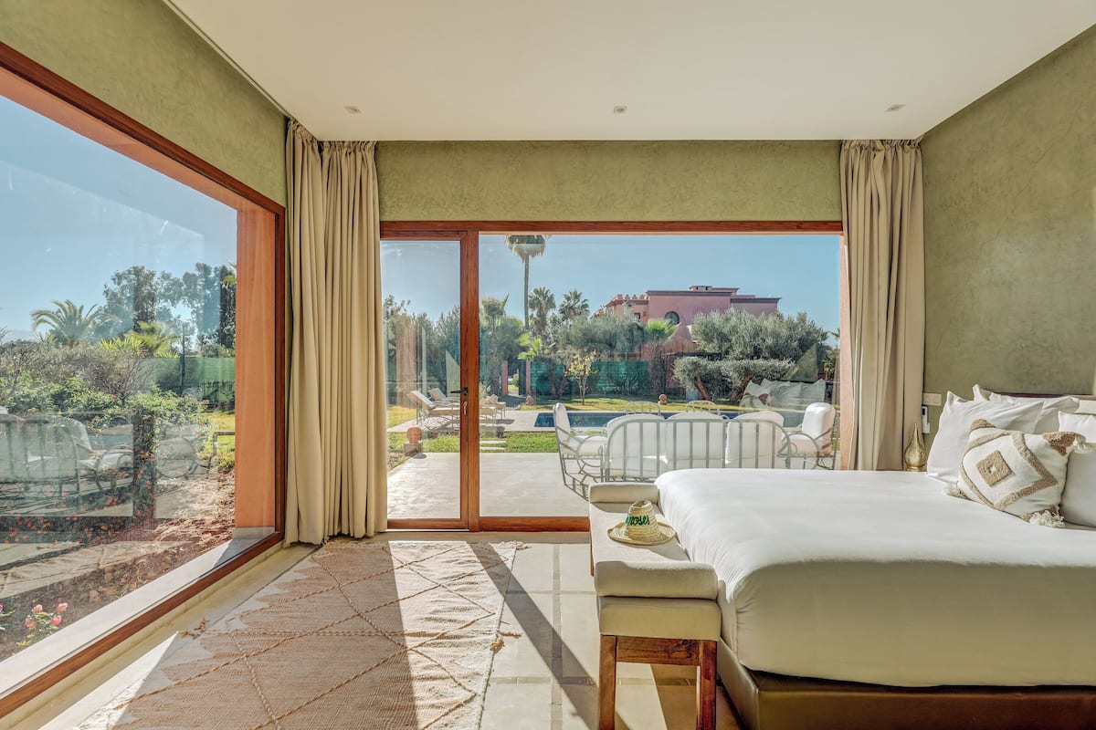 A bedroom with a large window, a bed, a bench, and a view of an outdoor area with a pool and chairs.