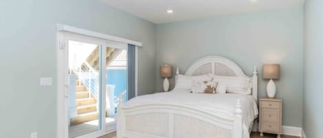 A bedroom with a white bed, two bedside lamps, a wooden nightstand, and a view of a blue building through a glass door.