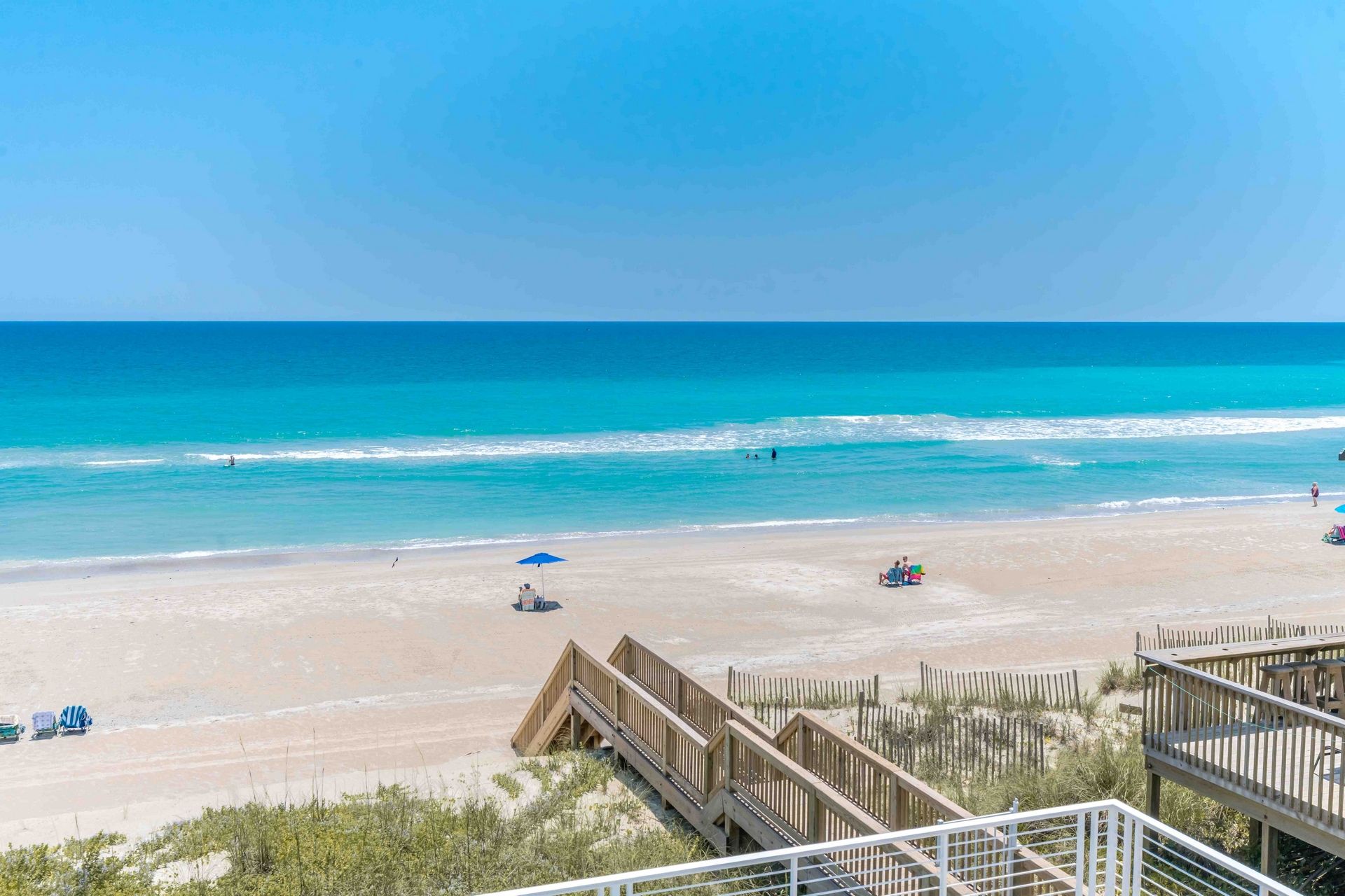 A beach with a wooden boardwalk leading to the shore, clear blue water, and a few people in the distance.