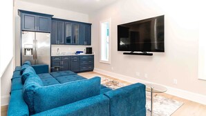 A modern living room with a sectional sofa, a flat-screen TV mounted on the wall, and a kitchen area with dark cabinets and a stainless steel refrigerator. - Ziggy's Place - New Luxury 8 BR Oceanfront Home; Pool & Elevator! (Surf City)