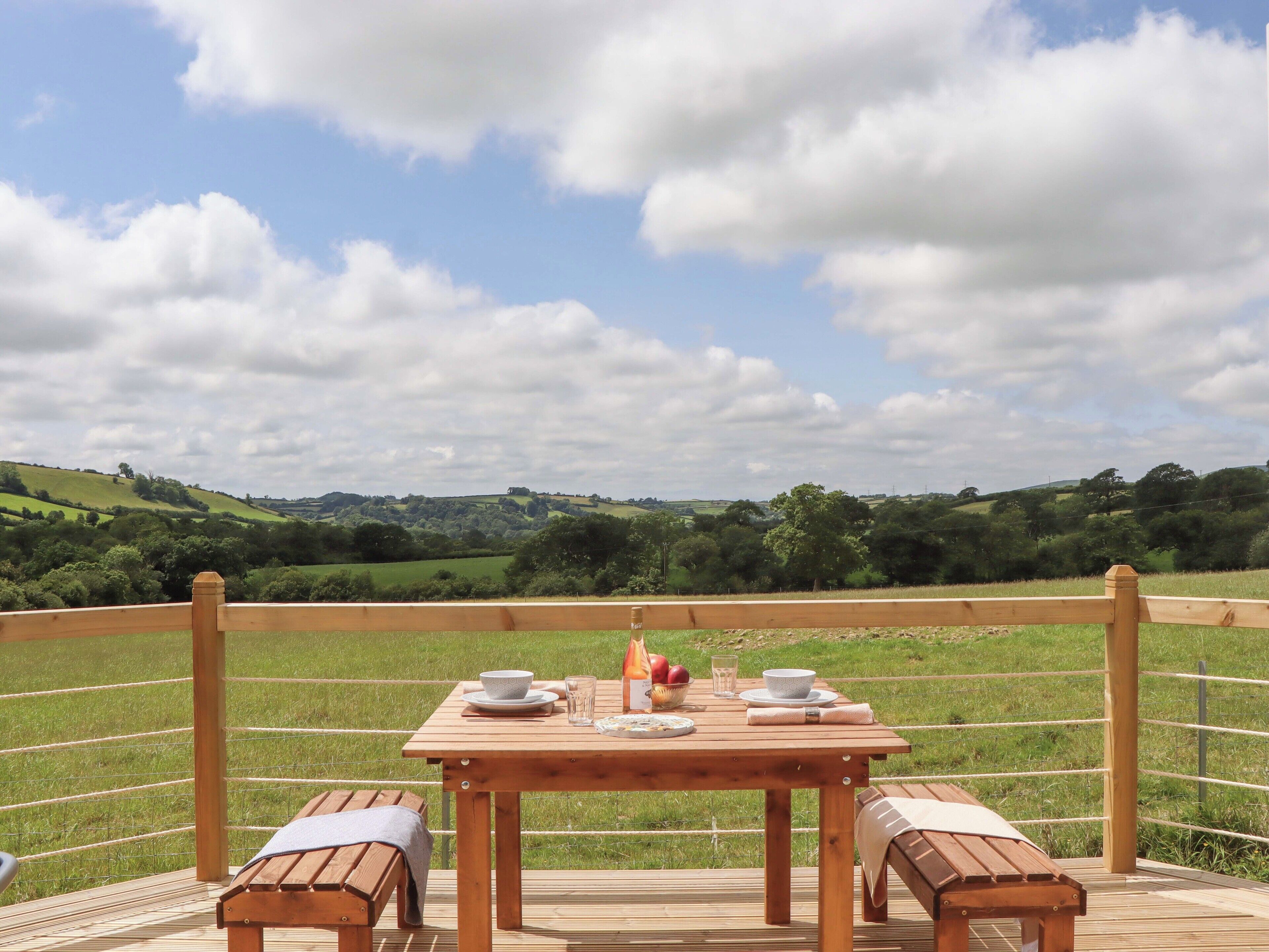Una terraza de madera con una mesa dispuesta para dos personas, con vistas a un campo cubierto de hierba y colinas ondulantes.
