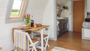 A cozy, well-lit room with a wooden dining table, chairs, and a small window. A kitchen area is visible in the background with modern appliances and a wooden door. - Under Oak Pod (Totnes)