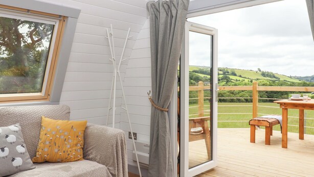 A cozy room with a sofa, a table, and a view of a green landscape through a sliding glass door. - Under Oak Pod (Totnes)