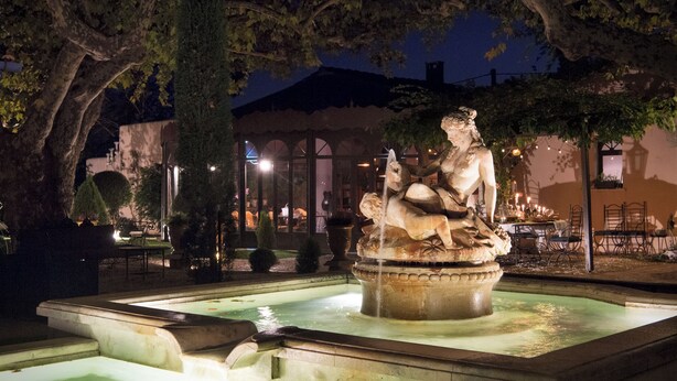 A nighttime outdoor fountain with a statue, surrounded by trees and a building with outdoor seating. - Domaine de l'Insoupçonnée (Noves)
