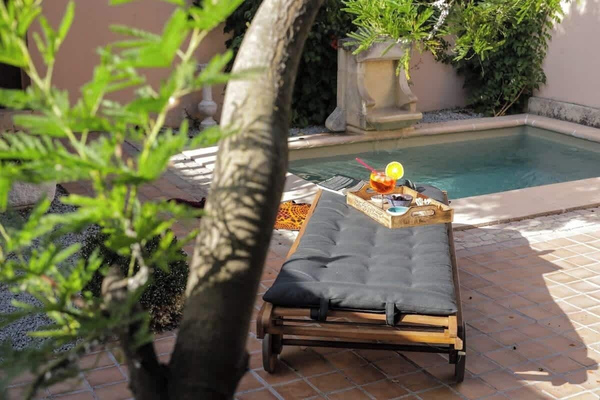 Una tumbona junto a una piscina, con una bebida y un libro, rodeada de follaje y un muro de piedra.