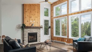 A spacious living room with a fireplace, large windows, and a ceiling fan. - Summer Getaway | Hot Tub, Kayaking & Mountain Biking (Saint-Côme)