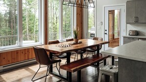 A spacious dining area with a wooden table and chairs, large windows offering a view of the forest, and a modern kitchen with cabinets and a toaster. - Summer Getaway | Hot Tub, Kayaking & Mountain Biking (Saint-Côme)