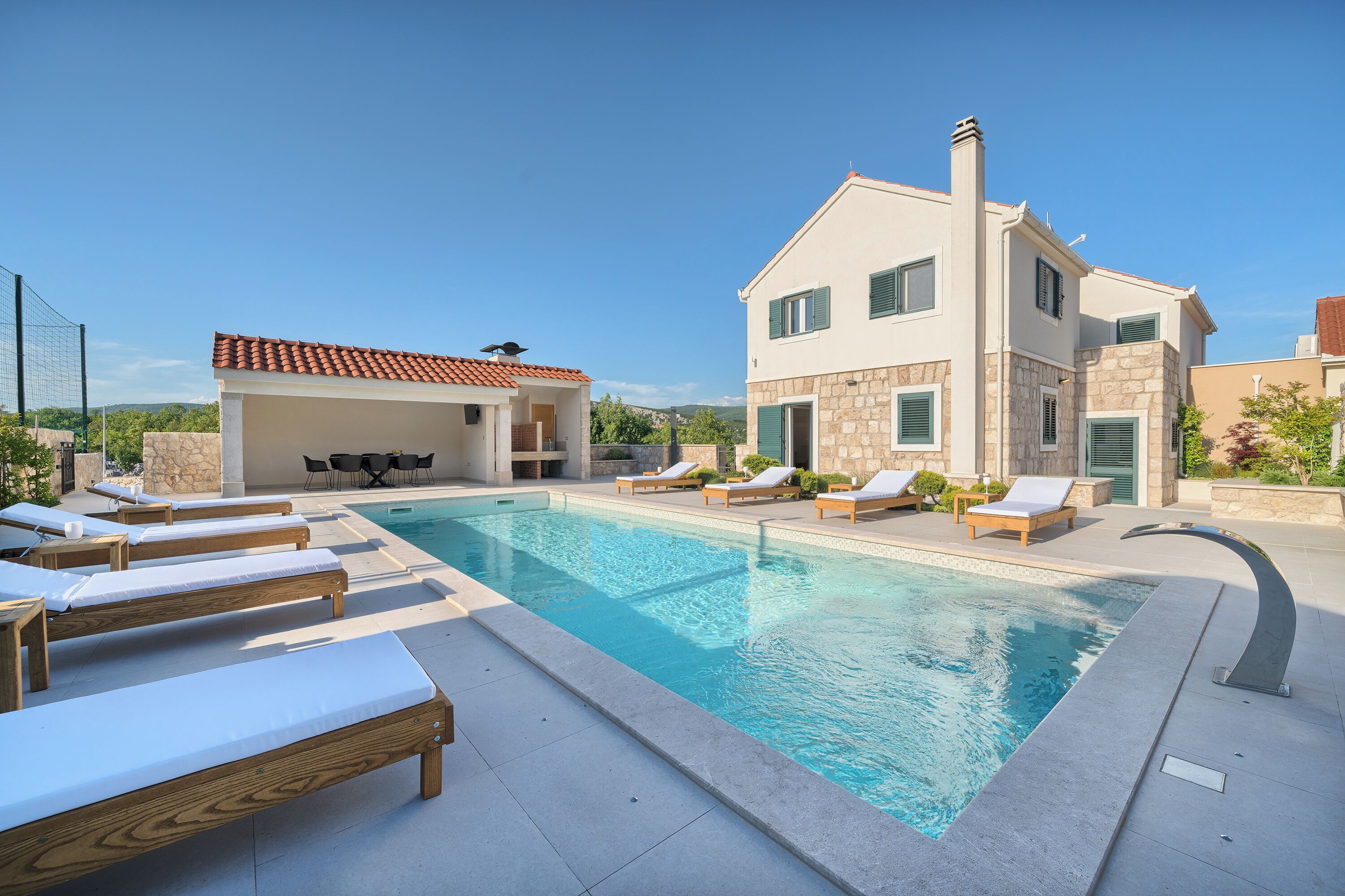 A swimming pool with lounge chairs, a covered area, and a two-story building with stone facade and green shutters.