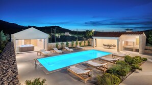 A swimming pool with lounge chairs, a hot tub, and a barbecue area, surrounded by a landscaped garden and a mountain backdrop.