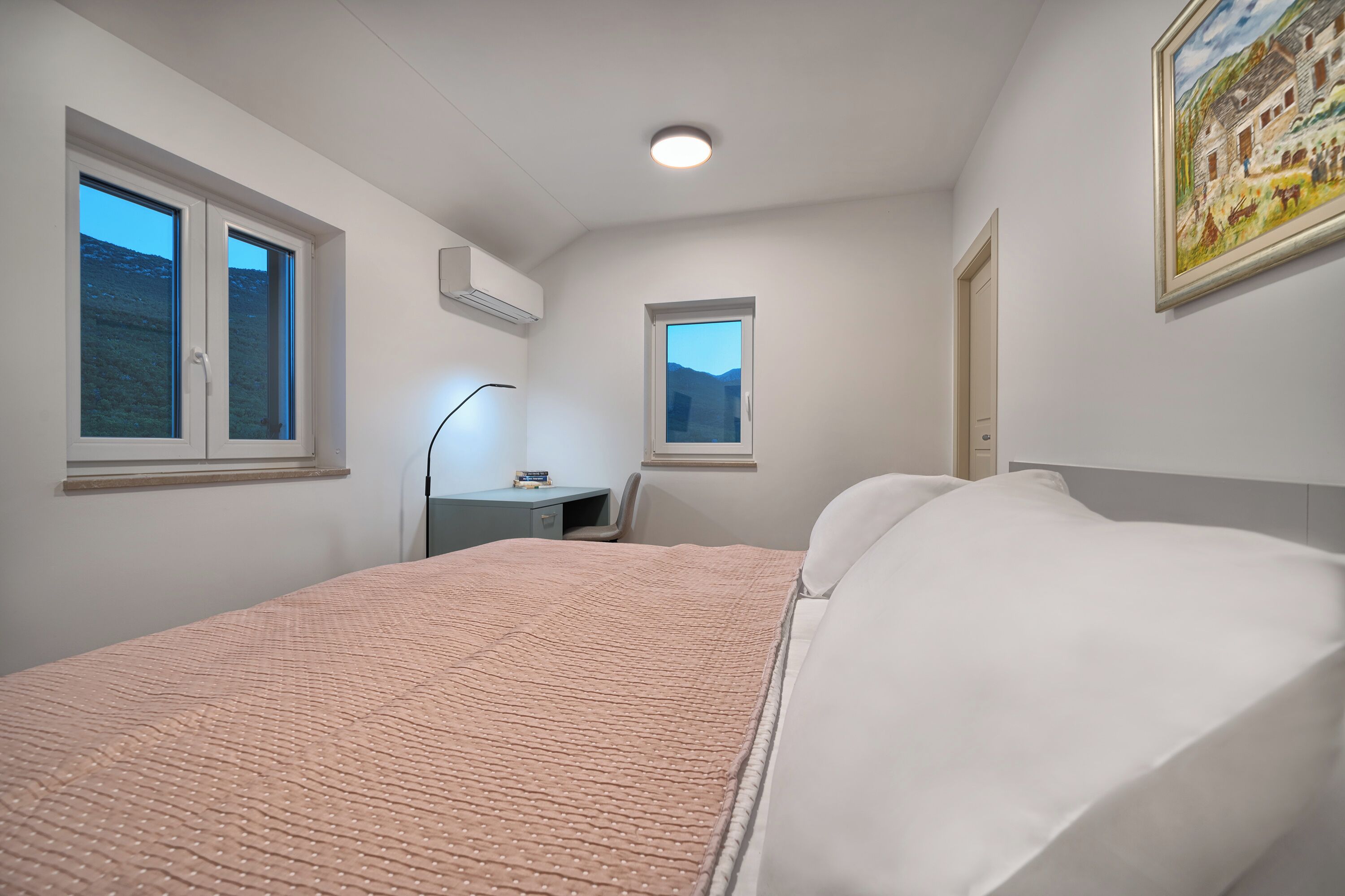 Un dormitorio con cama, dos ventanas, un escritorio y un cuadro en la pared.