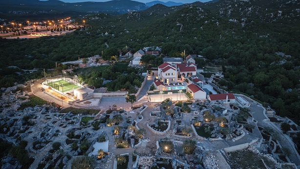 An aerial view of a resort with multiple buildings, a well-maintained garden, and a mountainous backdrop.
