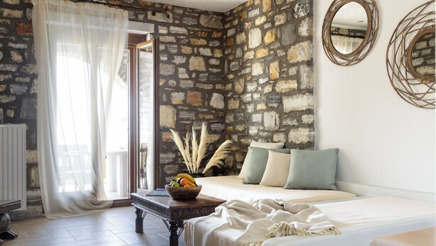 Une chambre avec un mur en pierre, un lit avec des oreillers, une table de chevet sur laquelle se trouve un bol de fruits, et une fenêtre ornée de rideaux légers.