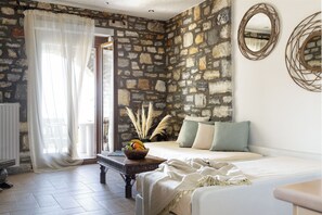 A bedroom with a stone wall, a bed with pillows, a bedside table with a bowl of fruit, and a window with sheer curtains. - Kaly Solana (Naxos)