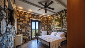 Une chambre avec des murs en pierre, un lit recouvert de draps blancs, un ventilateur de plafond et une vue sur l’extérieur, visible à travers une porte vitrée.