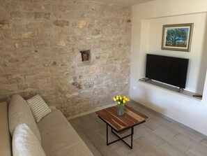 Un salon avec un mur en pierre, une table basse en bois sur laquelle est posé un vase rempli de fleurs jaunes, un canapé et un téléviseur à écran plat fixé au mur.