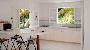 A kitchen with white cabinets, a white countertop, a microwave, a sink, and a window with a view of palm trees. - Résidence les Banians (Grand Gaube)