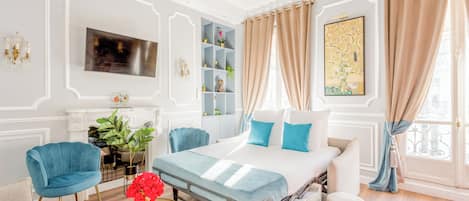 A hotel room with a bed, a TV, a bookshelf, a chandelier, and a window with curtains.