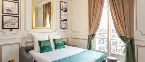 A hotel room with a bed, a large mirror, framed pictures on the wall, and a view of the city through the window.