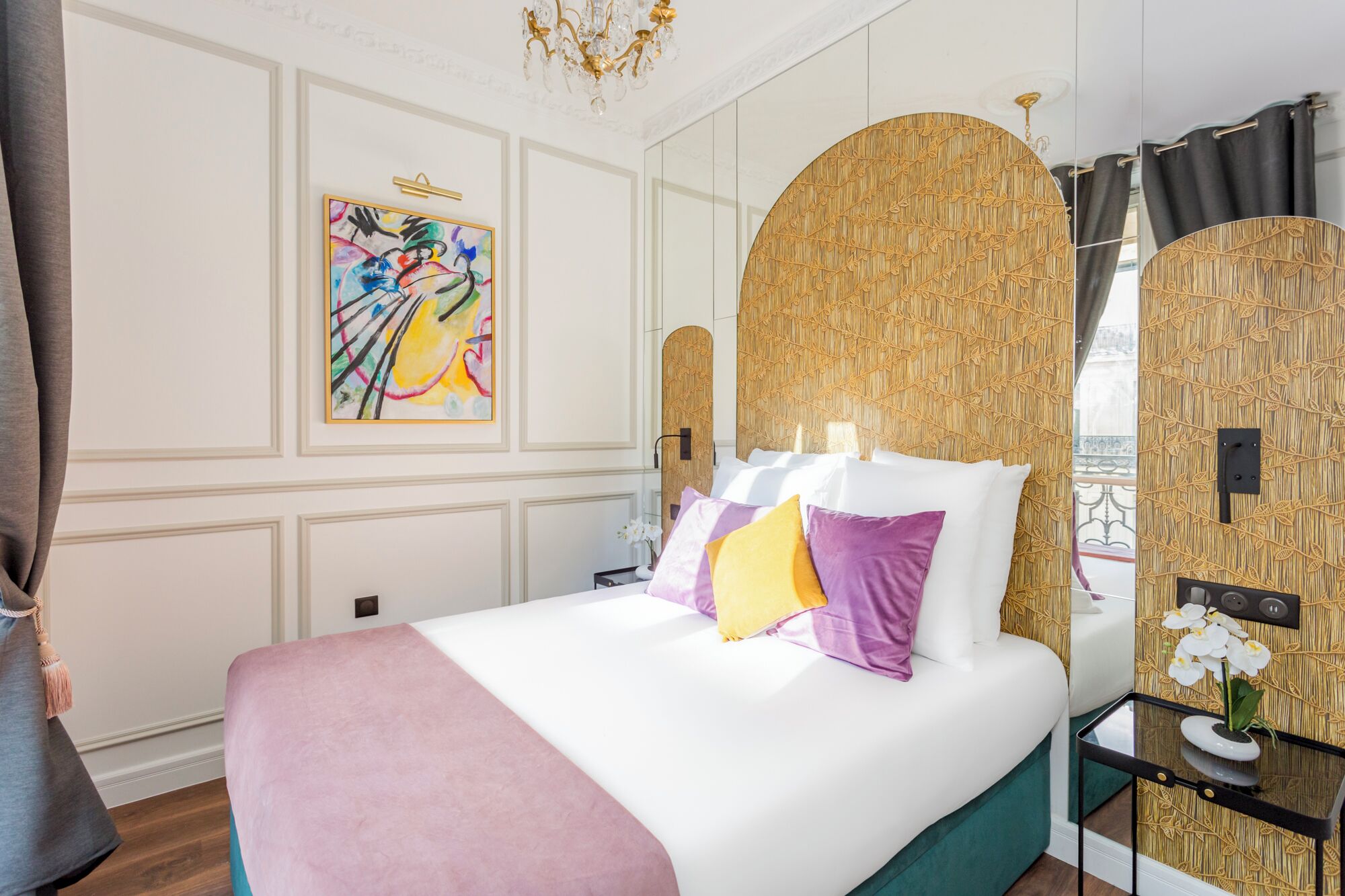 A modern hotel room with a large bed, a decorative wall panel, a chandelier, and a view through a window.