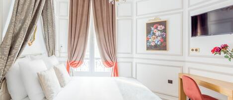 Un dormitorio con cama, cortinas, una araña, un escritorio, una silla, un televisor y un cuadro floral.