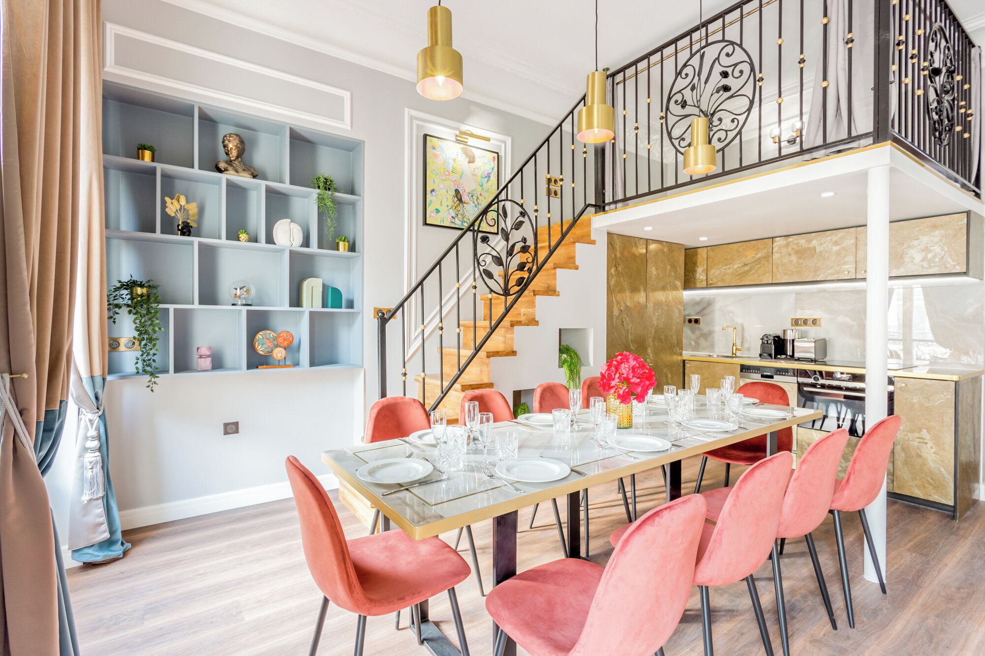 A dining area with a glass table set for six, red chairs, a staircase with a black railing, and a kitchen in the background.