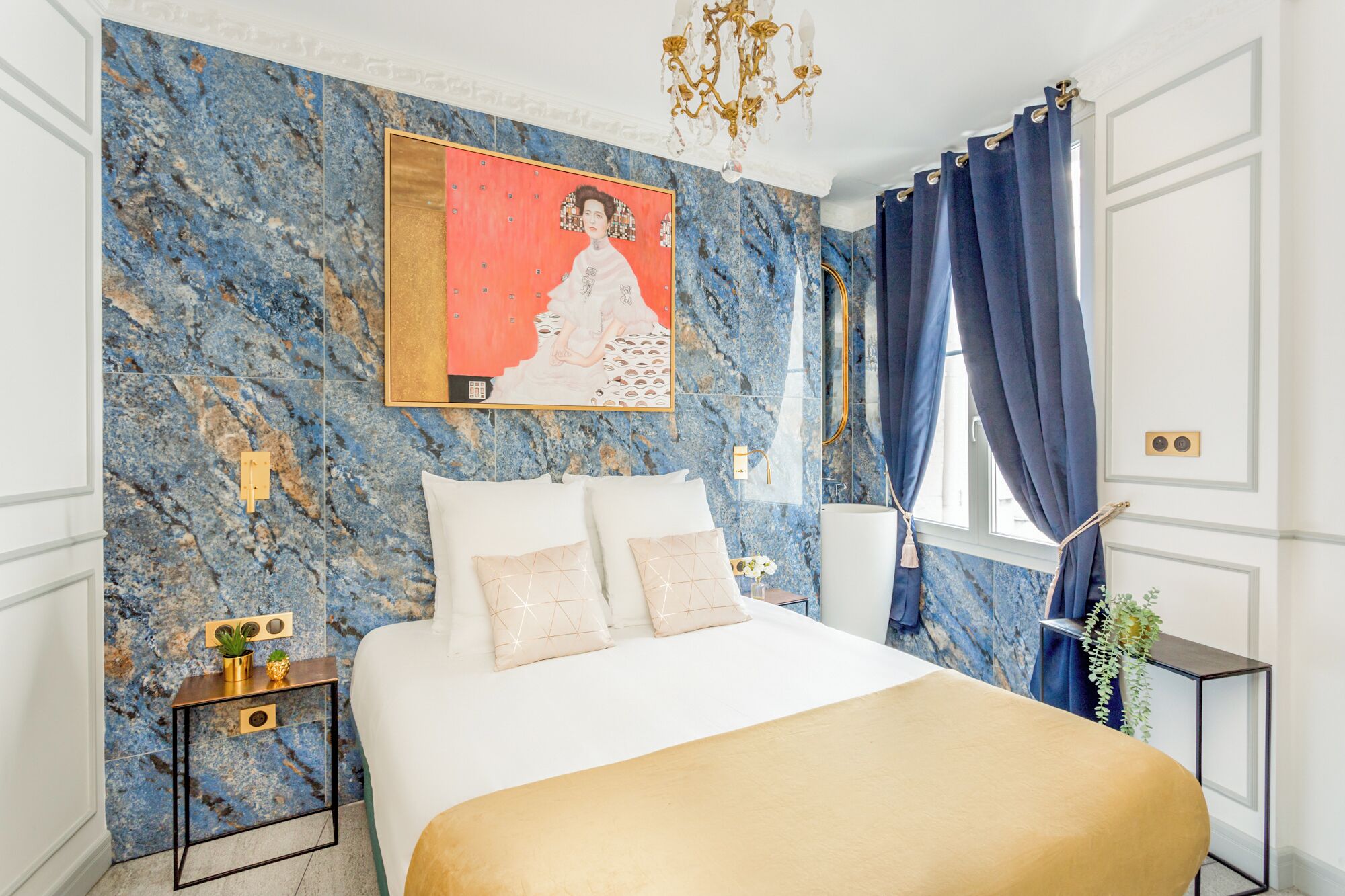 A bedroom with a bed, a nightstand, a painting, a chandelier, and a window with curtains.