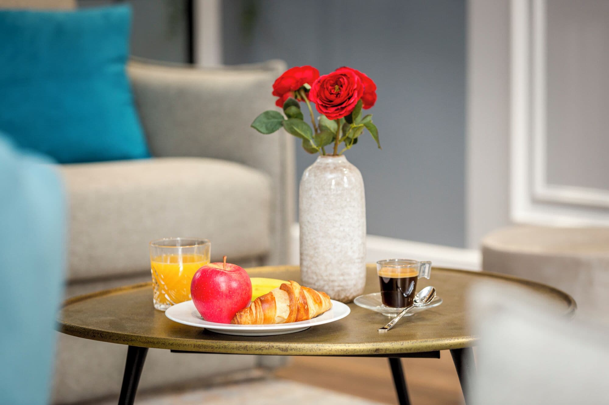A breakfast setting with a croissant, apple, orange juice, coffee, and a vase with red roses on a round table.