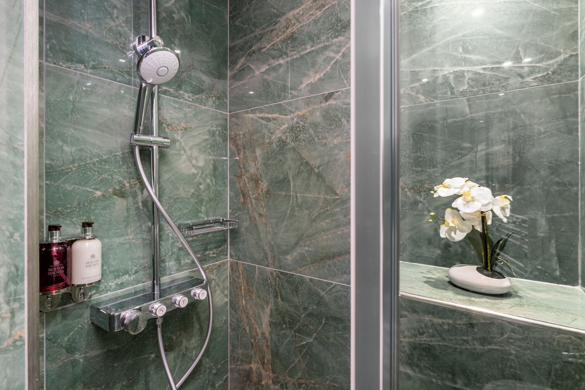 A shower with a handheld showerhead, a handheld shower attachment, and a wall-mounted control panel. There are two bottles on the wall, one labeled 'Molton Brown' and the other 'Molton Brown.' A potted orchid is visible on a shelf.