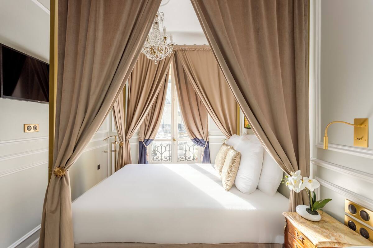 A bed with a canopy, a bedside table with a vase of flowers, and a view of the outside balcony.